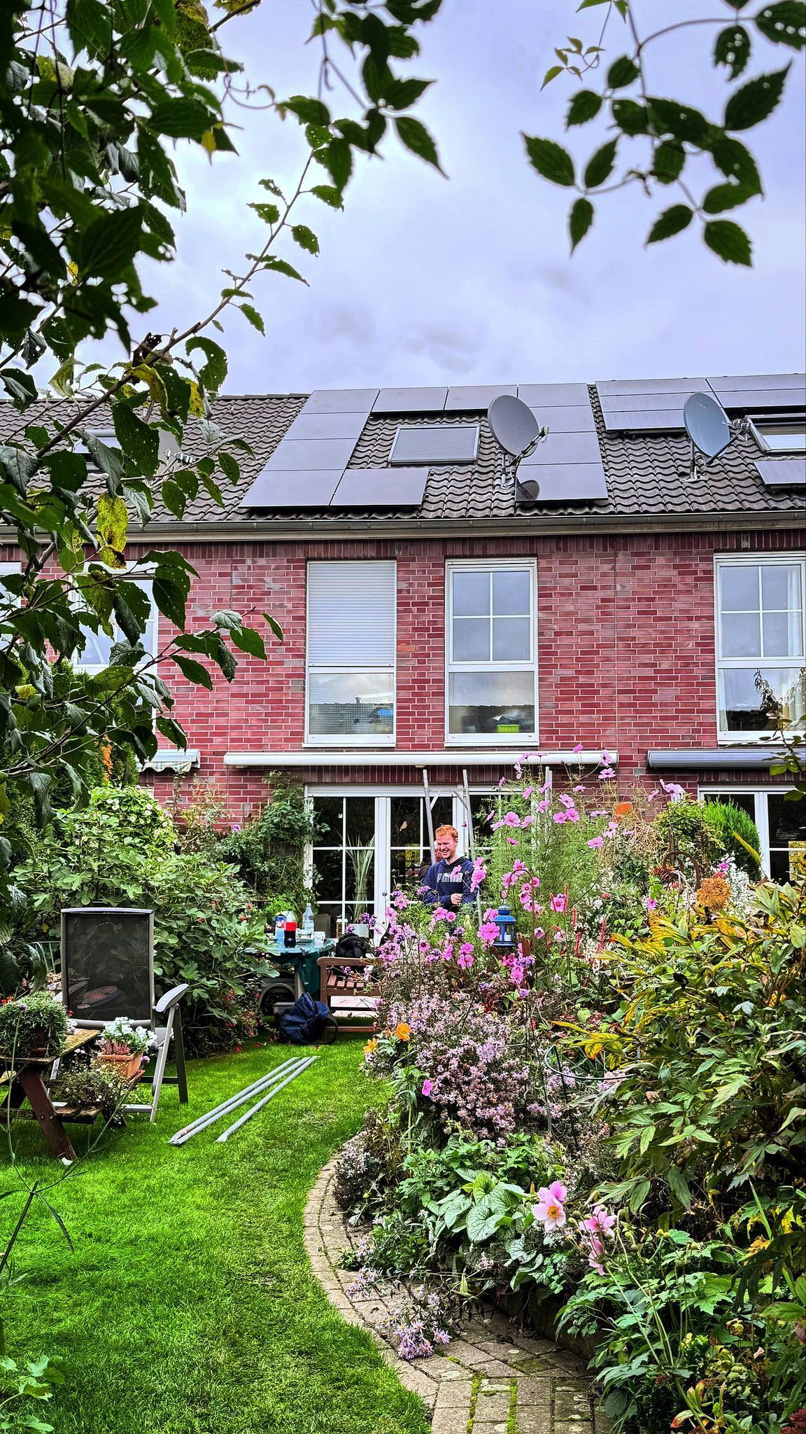 Ein rotes Backsteinhaus mit Solaranlage, umgeben von einem üppigen Garten. Eine Person steht in der Nähe der Terrasse und genießt die Natur.