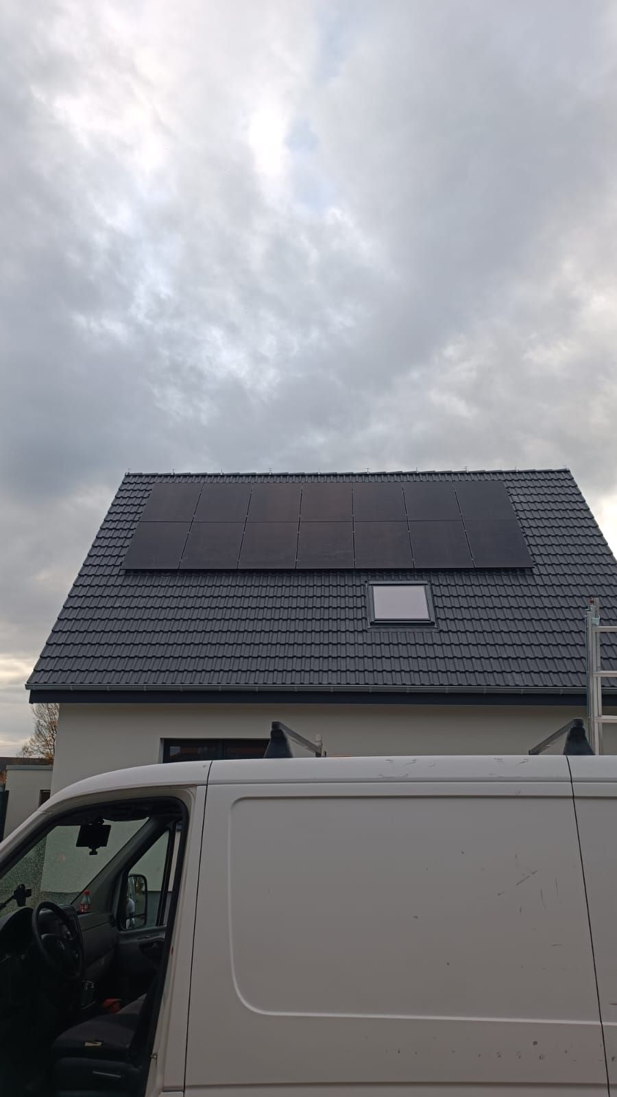 Solarpaneele auf einem Ziegeldach unter bewölktem Himmel, im Vordergrund ein weißer Lieferwagen.