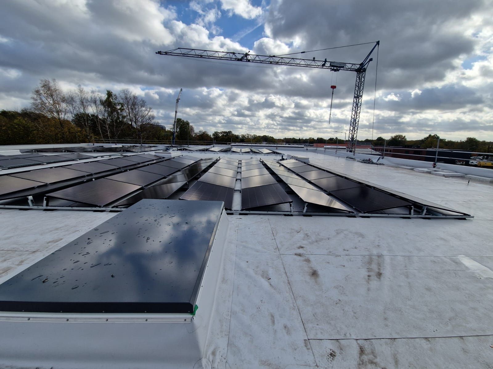 Auf einem weißen Dach sind Solarpaneele installiert, im Hintergrund ist unter einem bewölkten Himmel ein Baukran zu sehen.