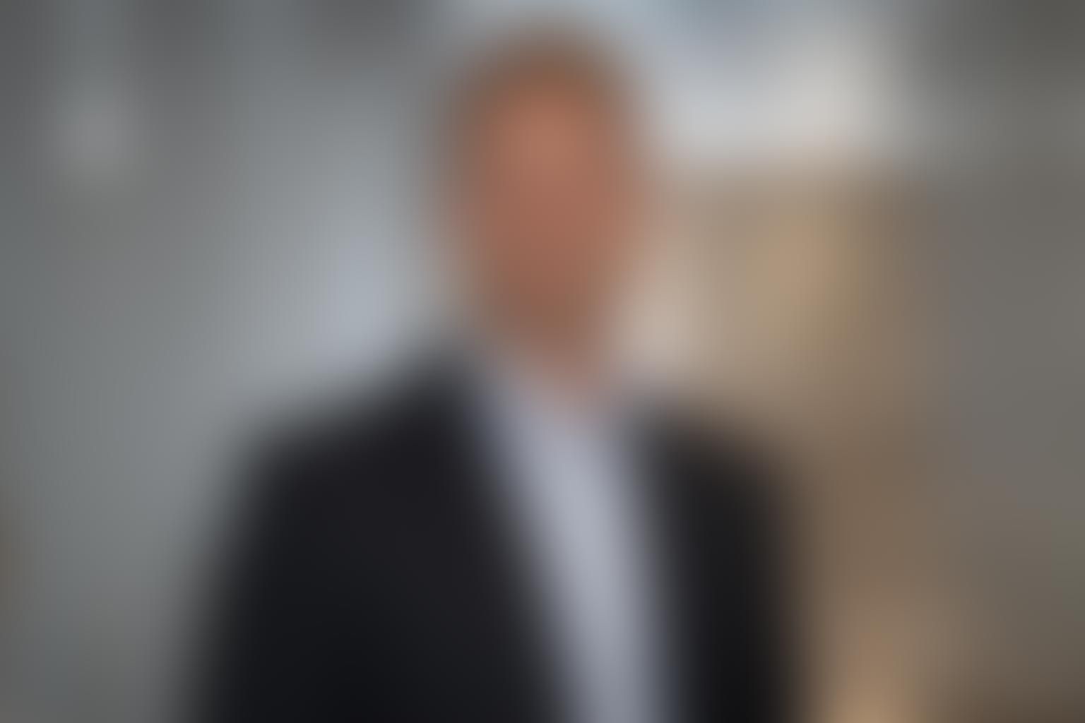 Mann in schwarzem Blazer und weißem Hemd, verschwommener Hintergrund.