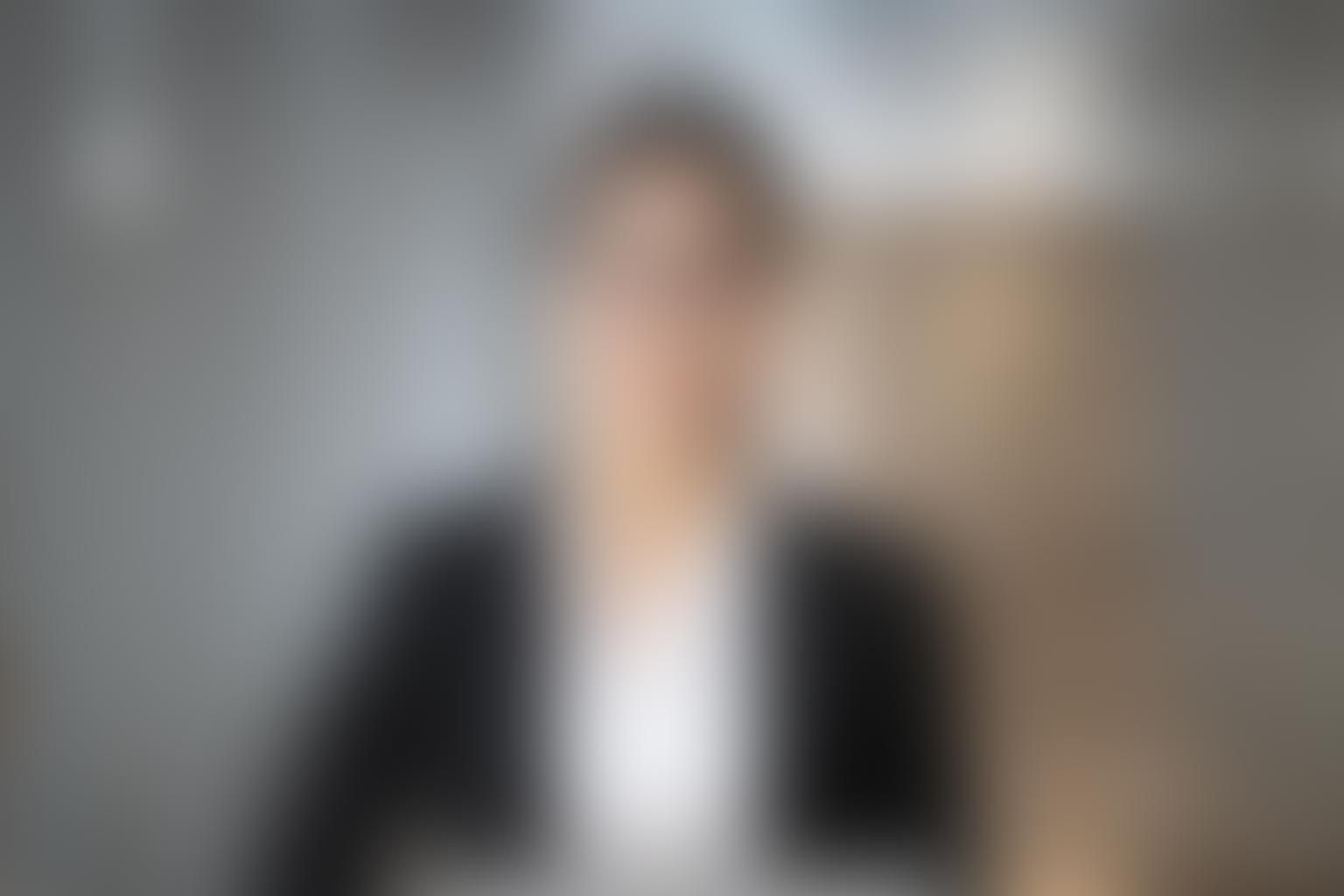 Person in schwarzer Jacke und weißem Hemd in einem Innenraum, unscharfer Hintergrund.