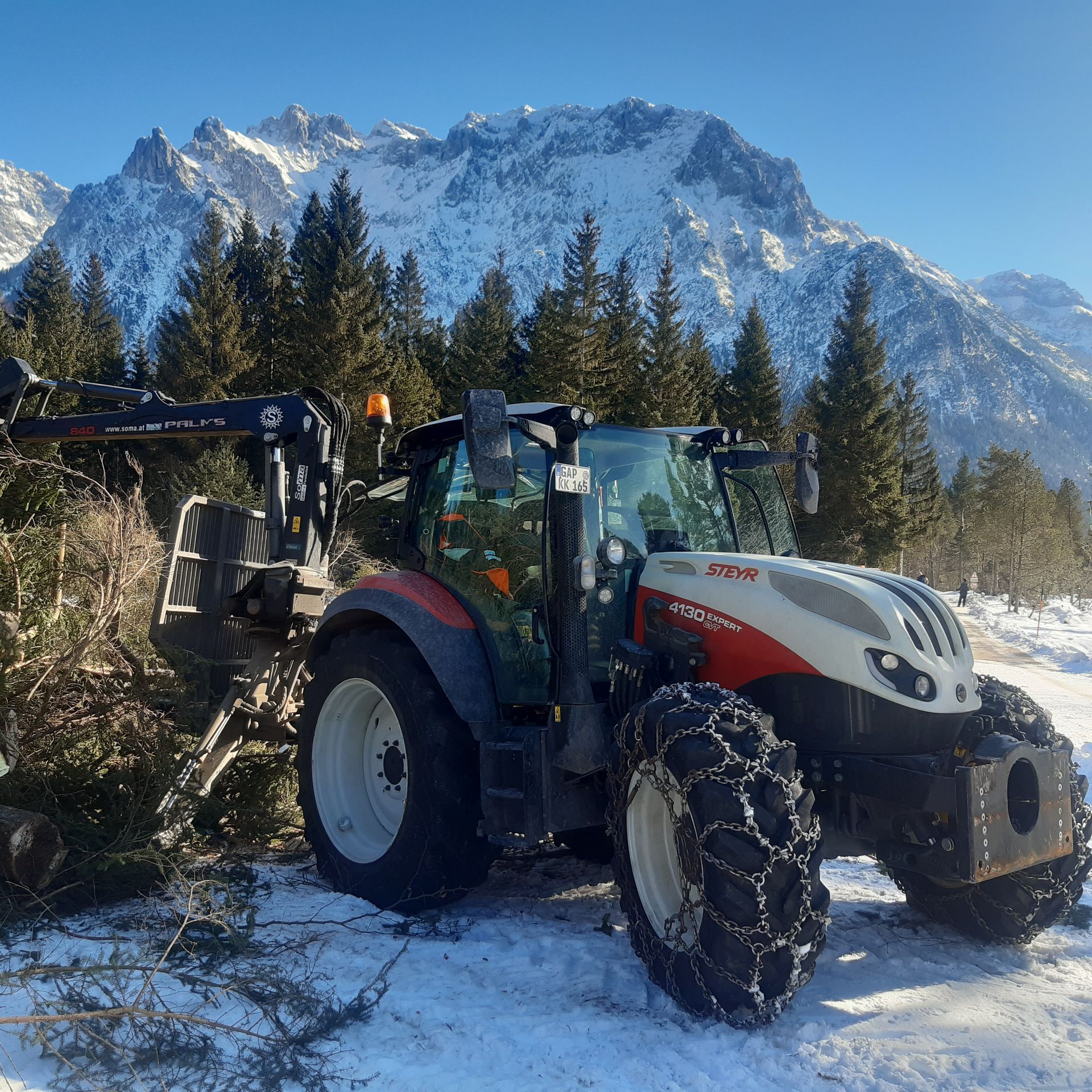 Nahaufnahme eines roten Traktors vor verschneiten Alpengipfeln im Sonnenschein.