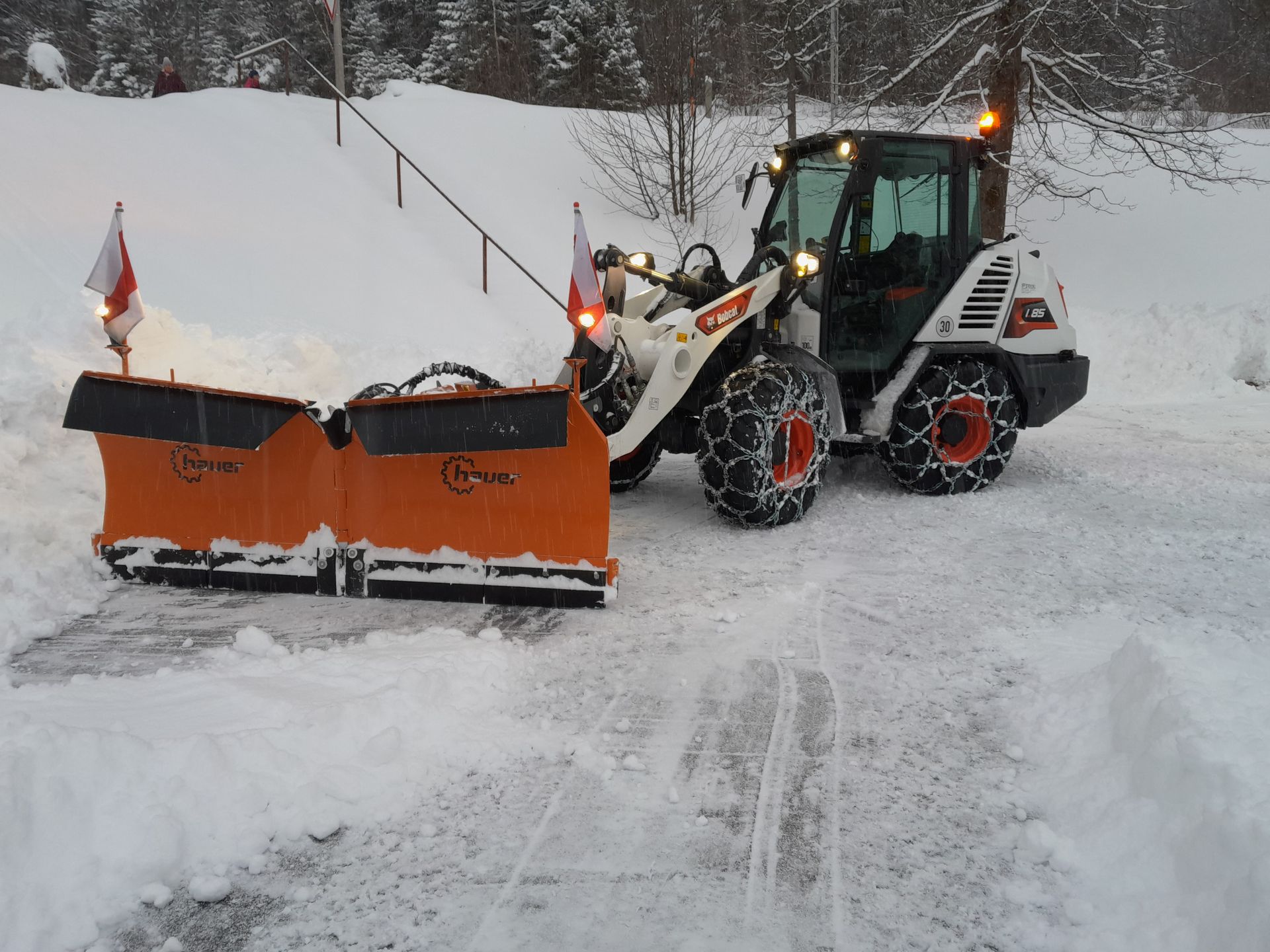 Kompaktlader mit großem Schneeschild räumt eine dicke Schneeschicht auf einem Parkplatz.