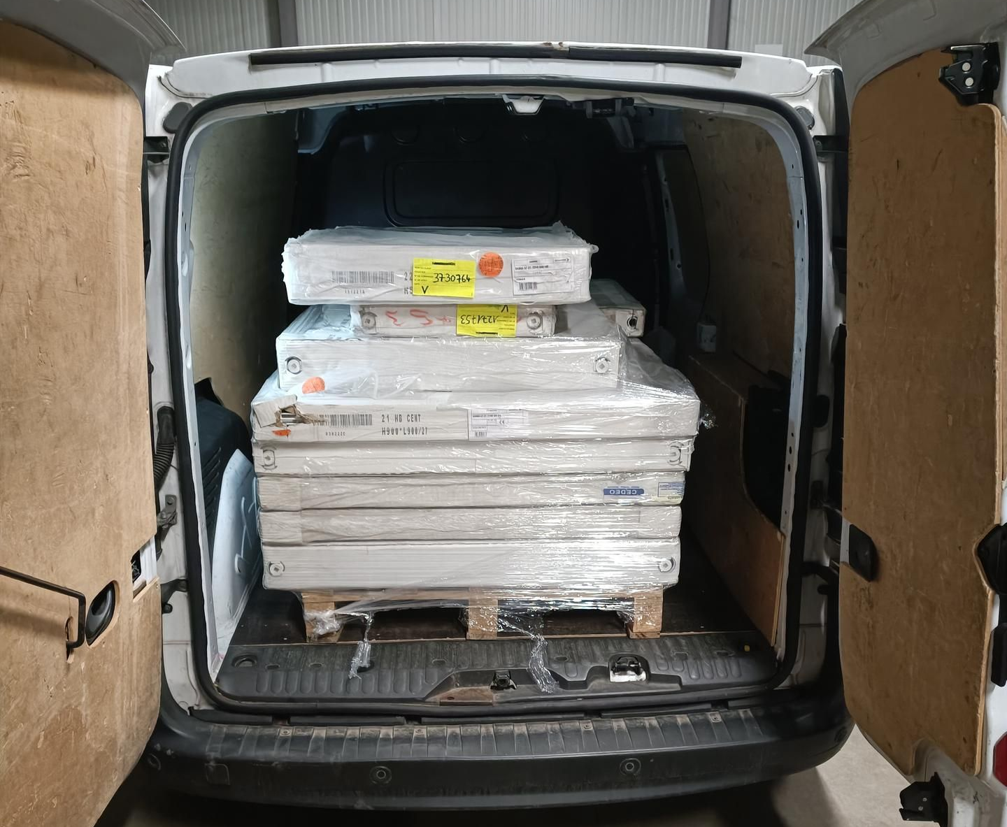 Fourgonnette blanche, portes ouvertes, chargée d'une pile de paquets rectangulaires emballés sur une palette.