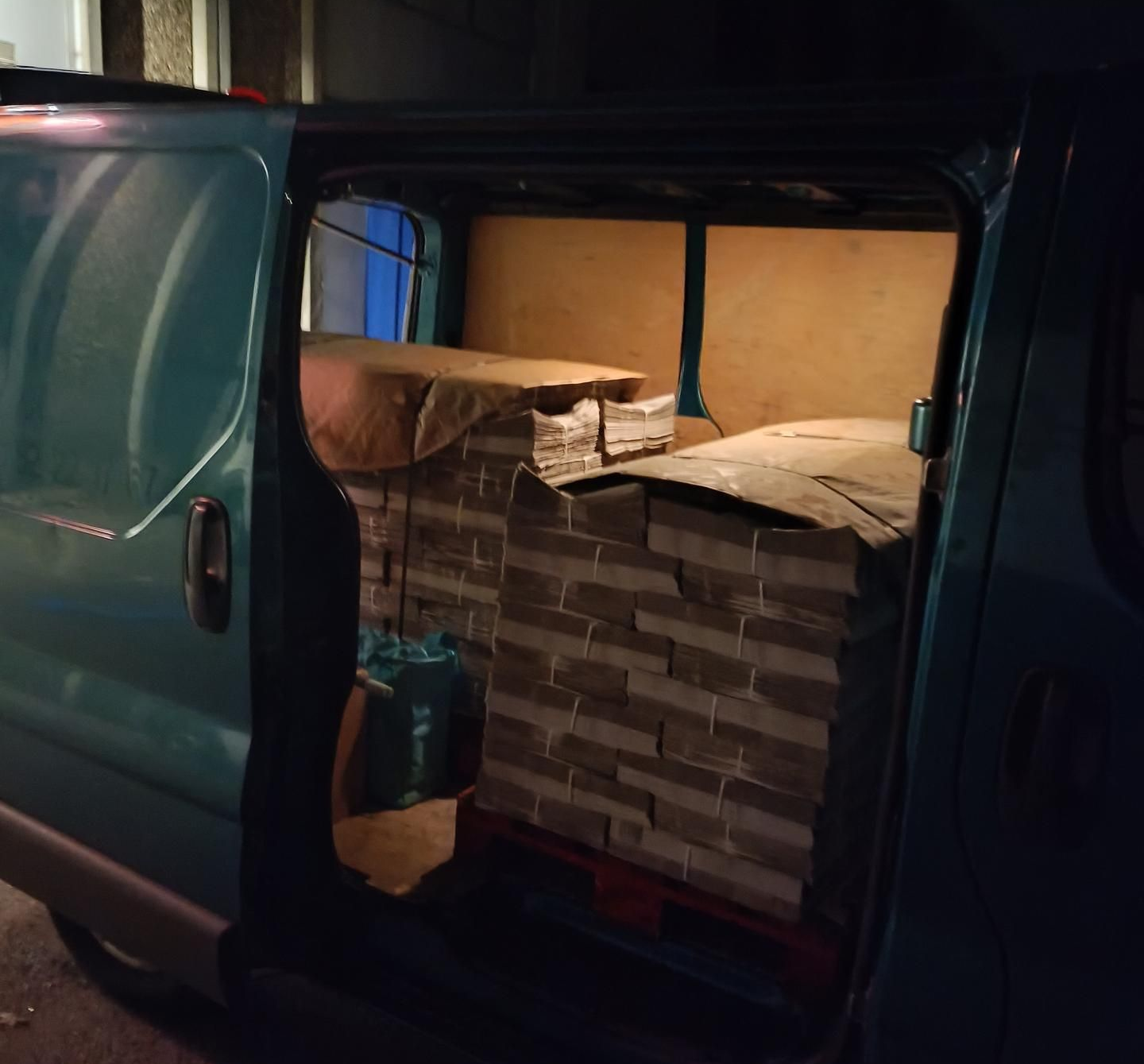 L'espace de chargement d'une camionnette turquoise est rempli de paquets empilés, emballés dans du papier kraft. La porte est ouverte.