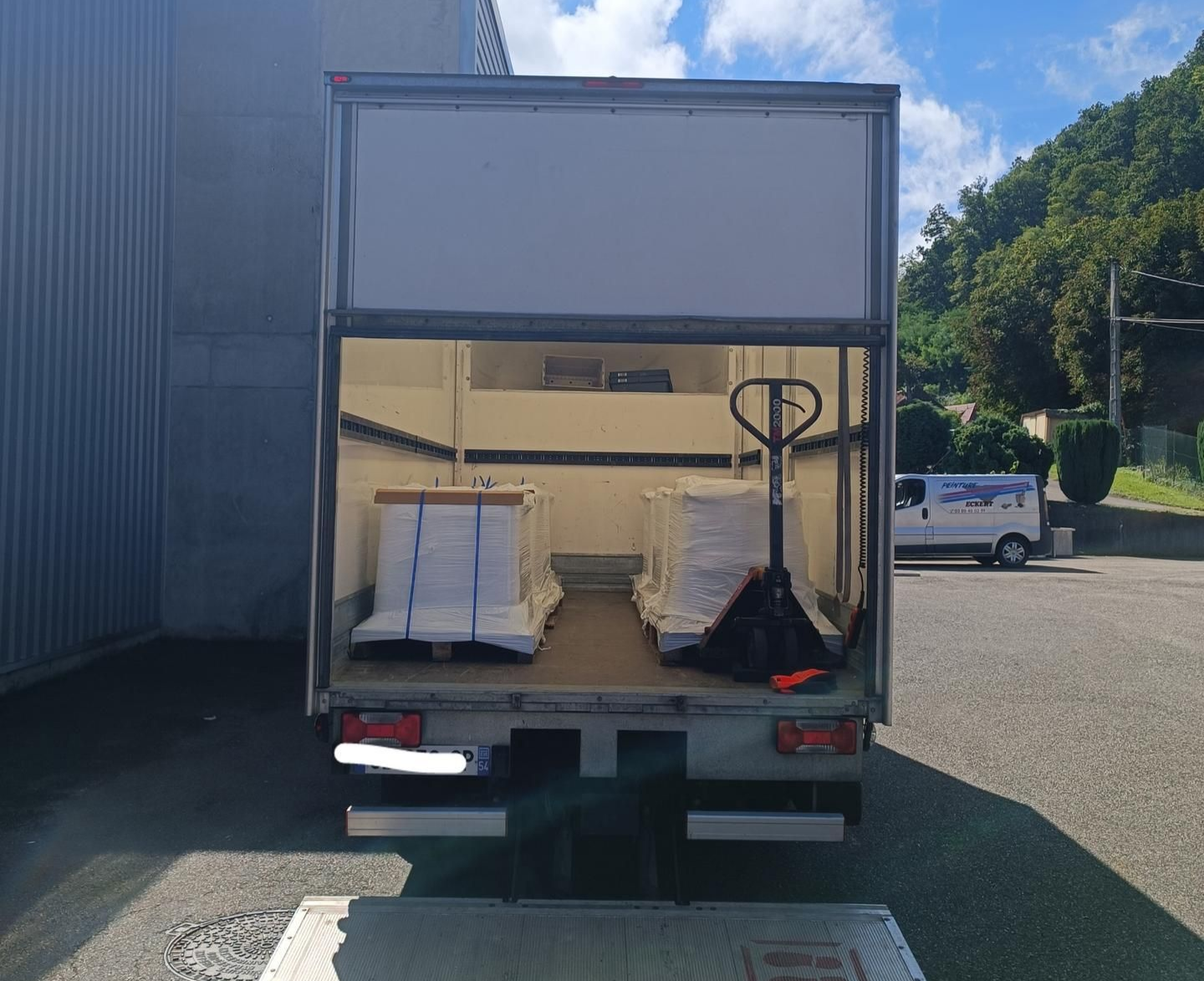 L'arrière ouvert d'un camion de livraison à hayon élévateur, chargé de palettes de marchandises, est stationné devant un bâtiment.