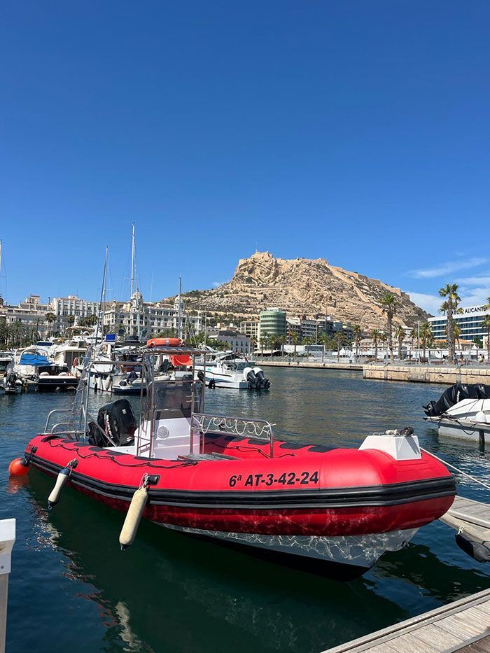Una lancha neumática roja atracada en un puerto deportivo con yates y una colina rocosa bajo un cielo azul despejado.
