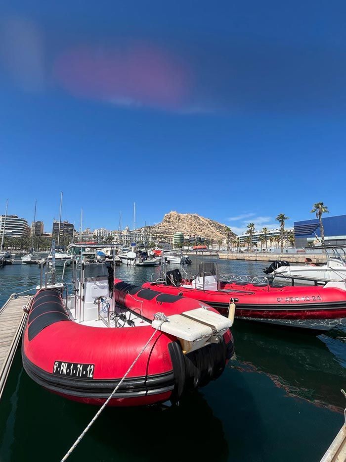 Botes inflables rojos atracados en un puerto deportivo bajo un cielo azul brillante, con una colina rocosa al fondo.