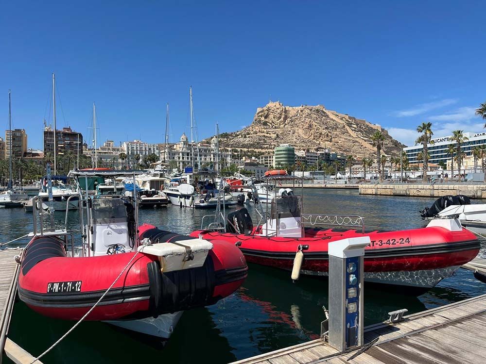 Botes inflables rojos amarrados en un puerto deportivo junto a veleros y una colina rocosa bajo un cielo azul.