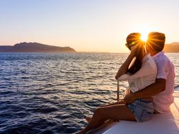 Una pareja se abraza en un barco al atardecer, con el sol reflejándose en el agua y una isla lejana en el horizonte.