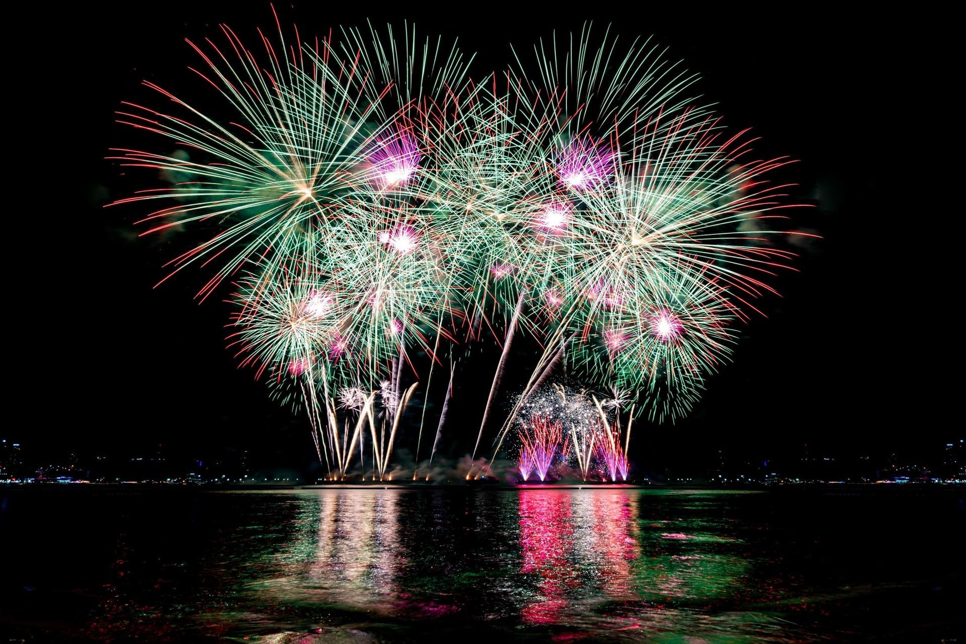 Fuegos artificiales de colores estallan sobre aguas oscuras, y sus reflejos brillan en la superficie.
