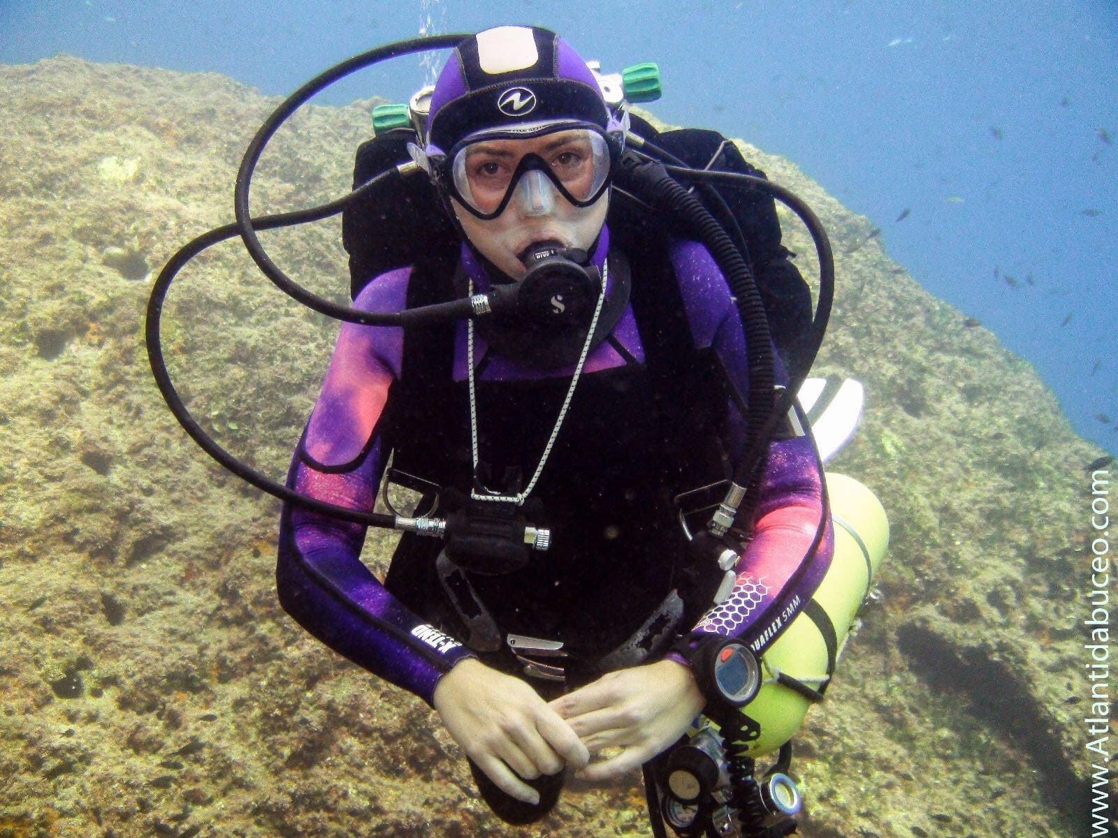 Buzo con traje morado nadando sobre un fondo marino rocoso con arrecife de coral en aguas azules.