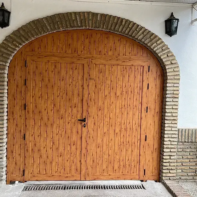 Puerta doble de madera arqueada con detalles de ladrillo y faroles negros sobre una pared blanca.