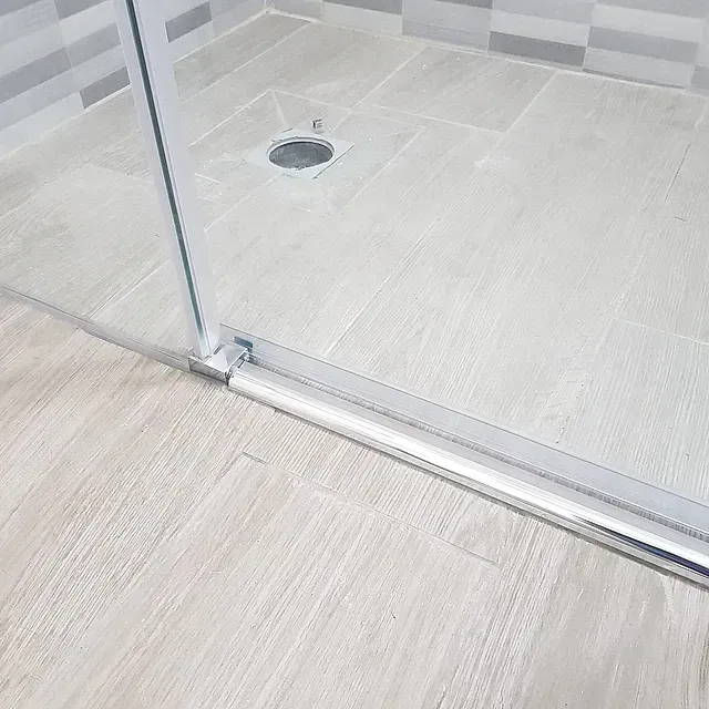 Puerta de ducha de cristal con riel cromado y desagüe en el suelo en un baño con azulejos claros.