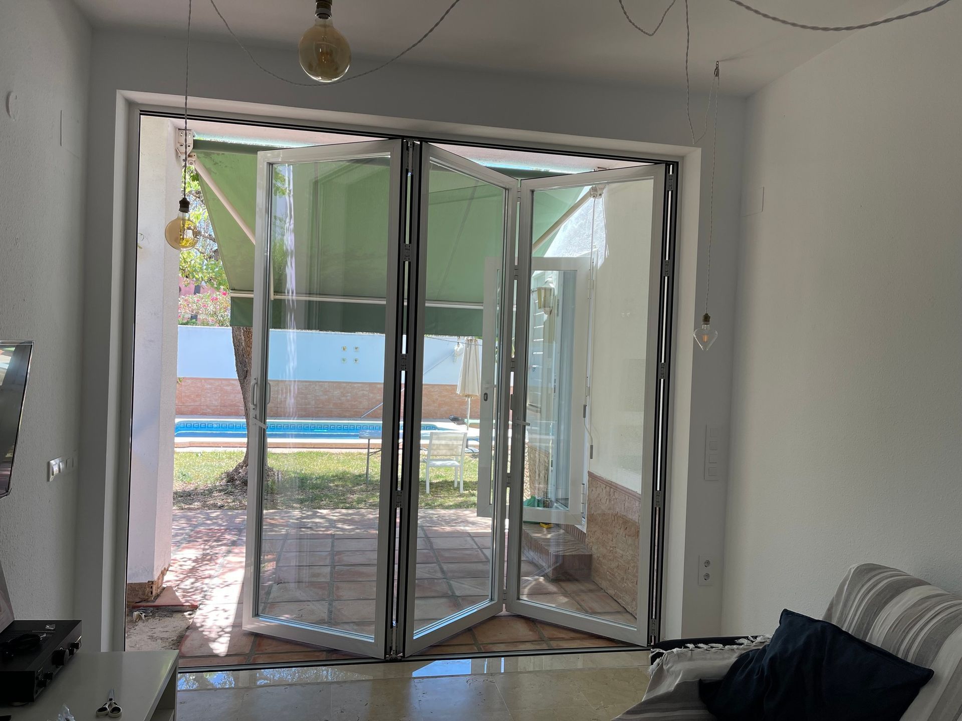 Puertas de cristal parcialmente abiertas que dan a un luminoso patio y jardín, con un sofá a la derecha.
