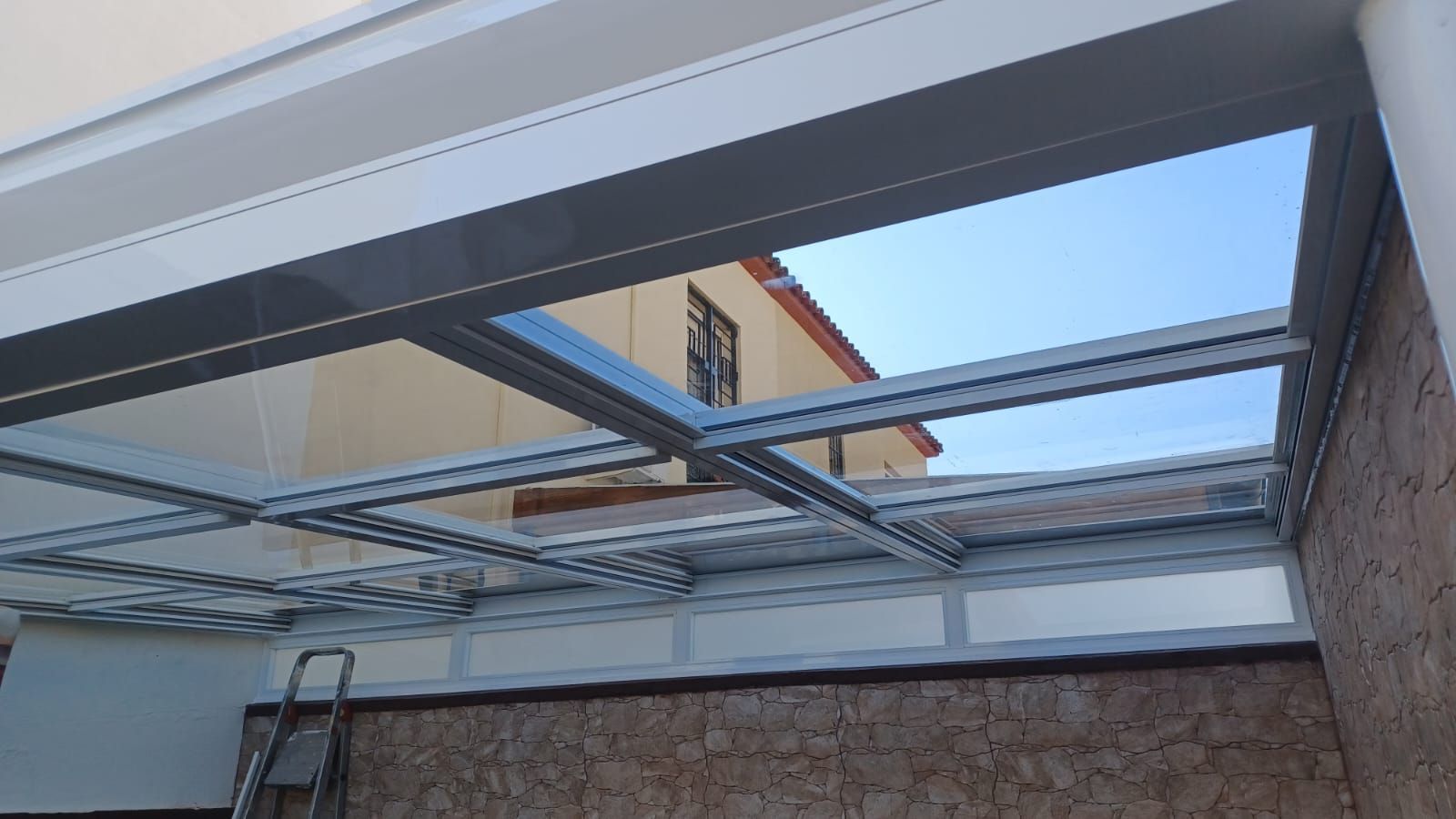 Techo con tragaluz parcialmente abierto sobre un patio, con vigas metálicas y cielo azul a la vista.