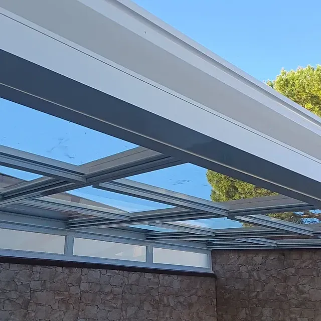 Techo de cristal con estructura metálica blanca sobre un muro de piedra, visto contra un cielo azul.