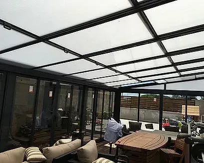 Patio exterior cubierto con paredes de cristal, una mesa, cojines y una persona con camisa blanca cerca de la zona de estar.