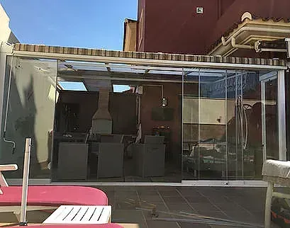 Patio exterior con paredes de cristal, sillas y mesas oscuras bajo un toldo blanco junto a un edificio.