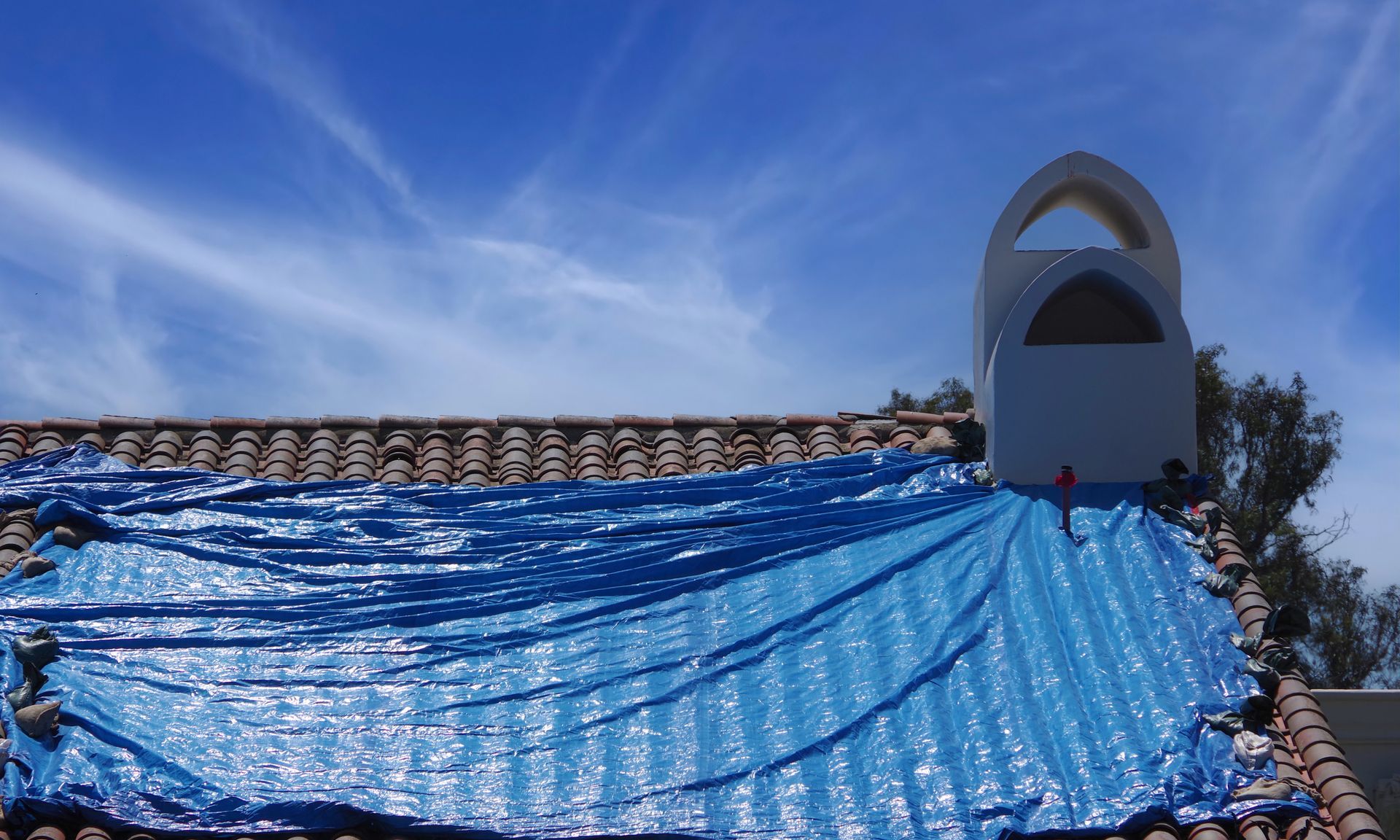 Une bâche bleue recouvre une partie endommagée d'un toit de tuiles, à côté d'une cheminée blanche en arc de cercle