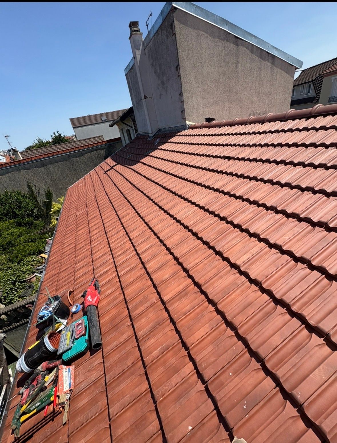Un toit de tuiles rouges sous un ciel bleu, avec une cheminée, une ventilation de toit et des outils de construction posés sur les tuiles