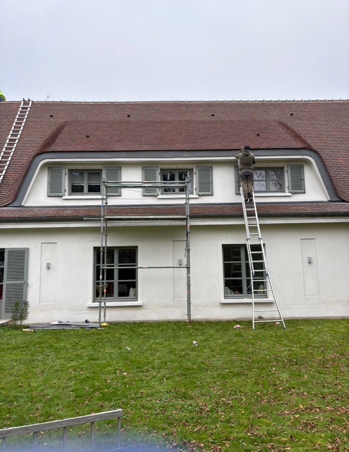 Une personne sur une échelle répare le toit d'une maison blanche à lucarnes, un échafaudage est présent à côté.