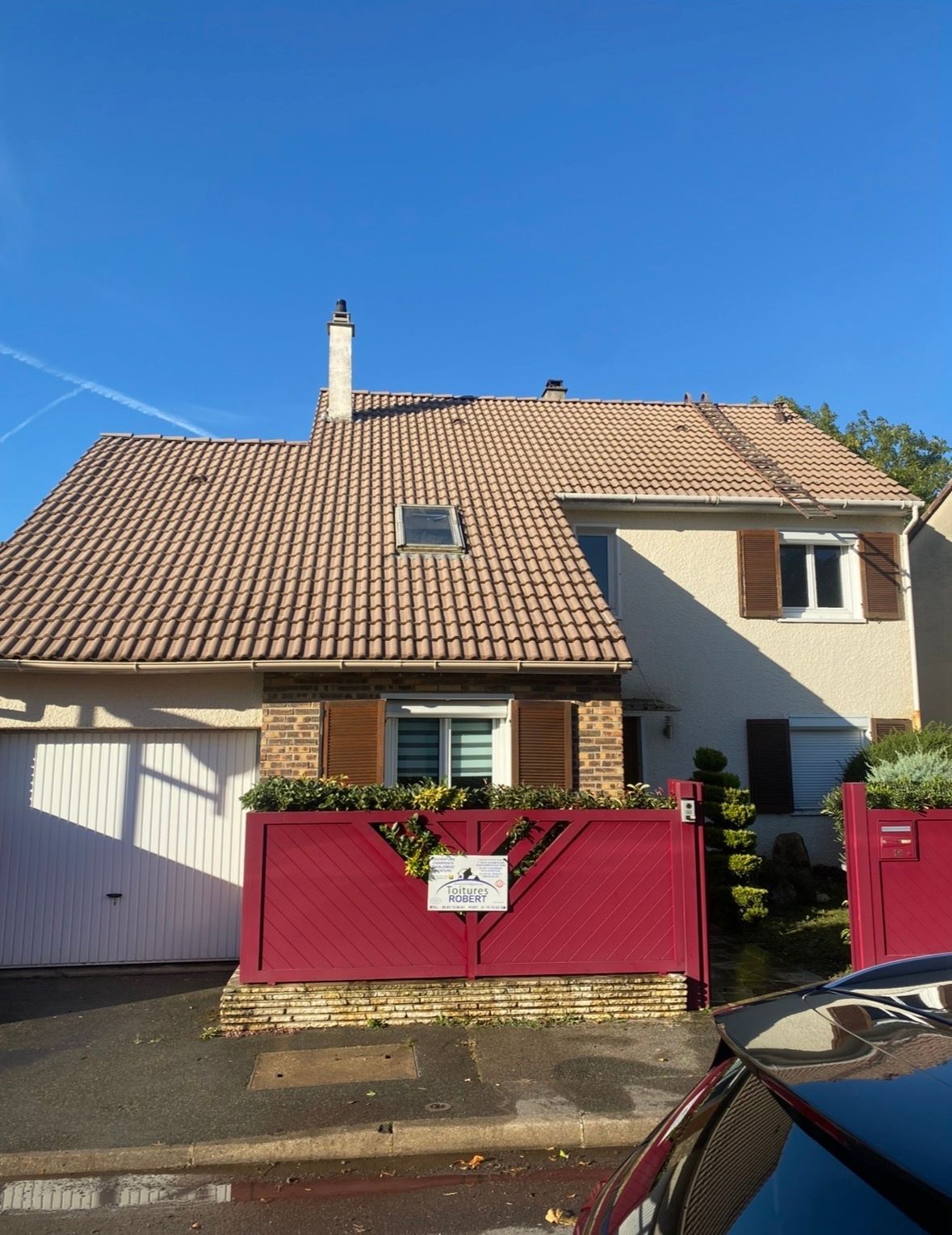 Une maison claire avec un étage avec un toit de tuiles brunes, un garage blanc et une clôture en bois rouge