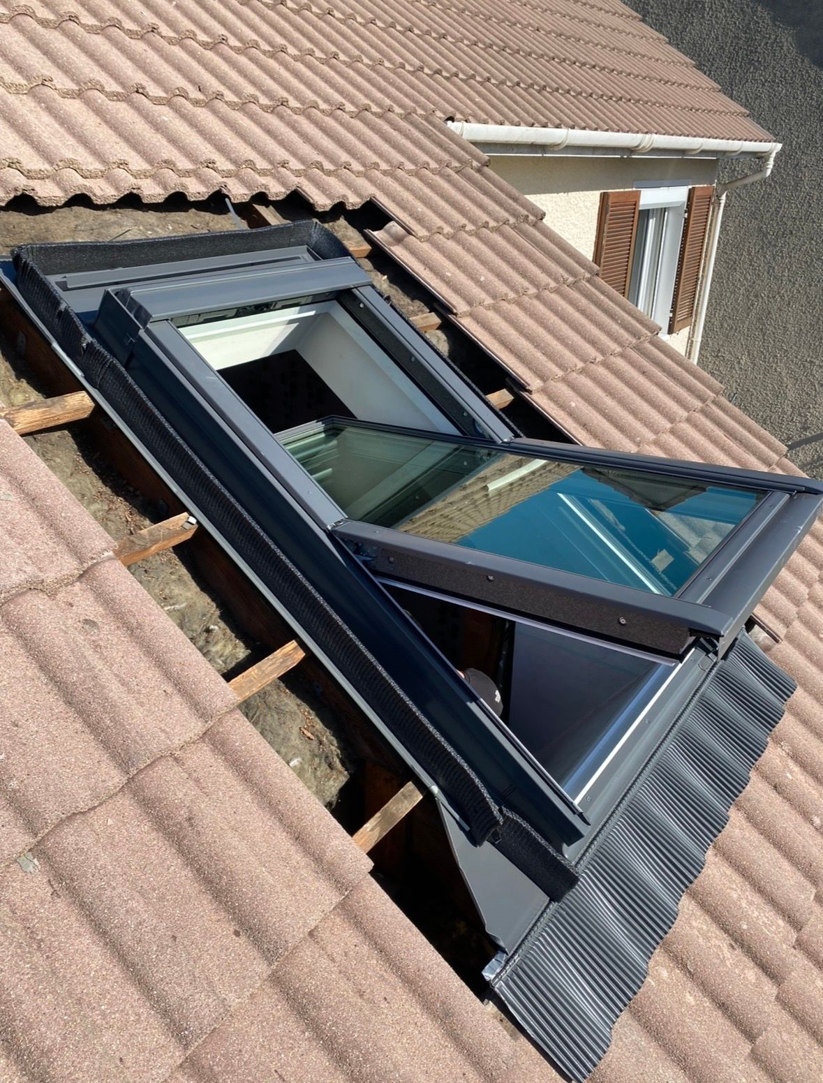 Une fenêtre de toit partiellement installée sur un toit de tuiles avec des liteaux en bois apparents et des tuiles