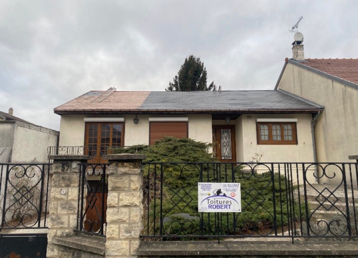 Une maison de plain-pied avec un toit gris partiellement nettoyé, une cour avant avec un buisson et une clôture en fer forgé