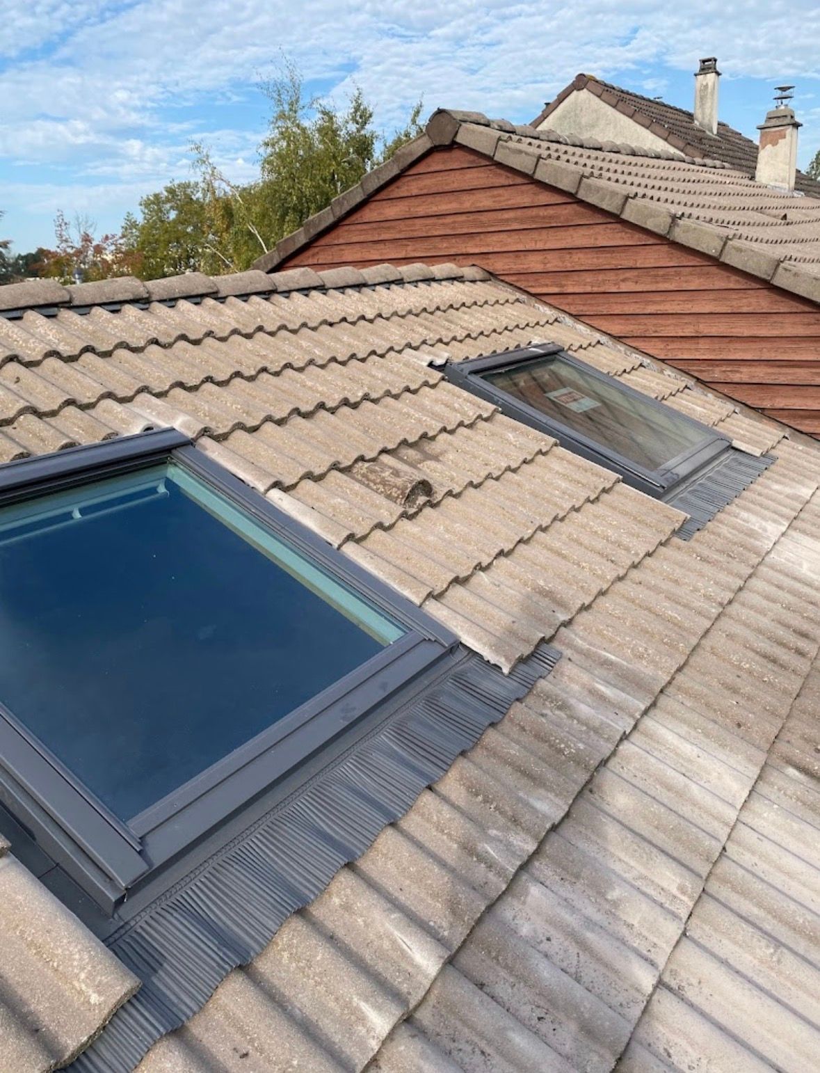 Deux fenêtres de toit installées sur un toit en pente gris, recouvert de tuiles, près d'une structure en briques