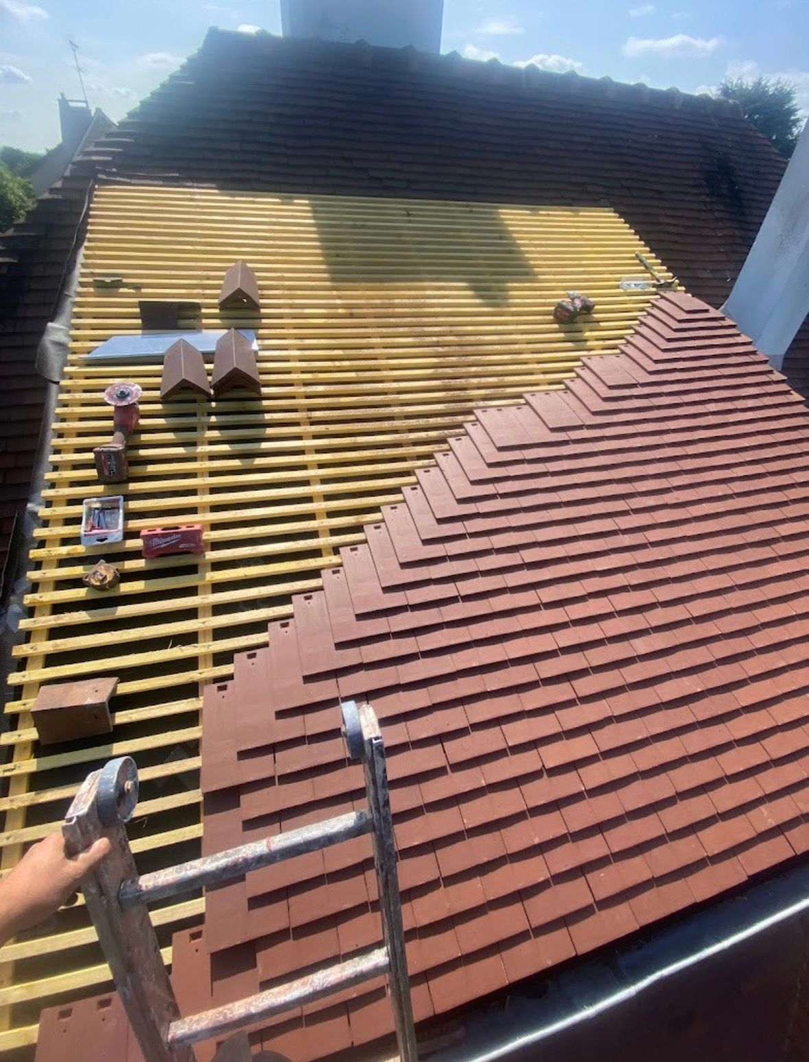 Vue depuis une échelle montrant un toit en construction, une partie étant recouverte de tuiles rouges et l'autre de liteaux apparents