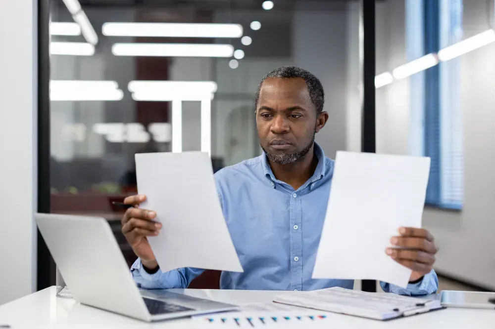 Um homem está sentado em uma mesa segurando dois pedaços de papel na frente de um laptop.