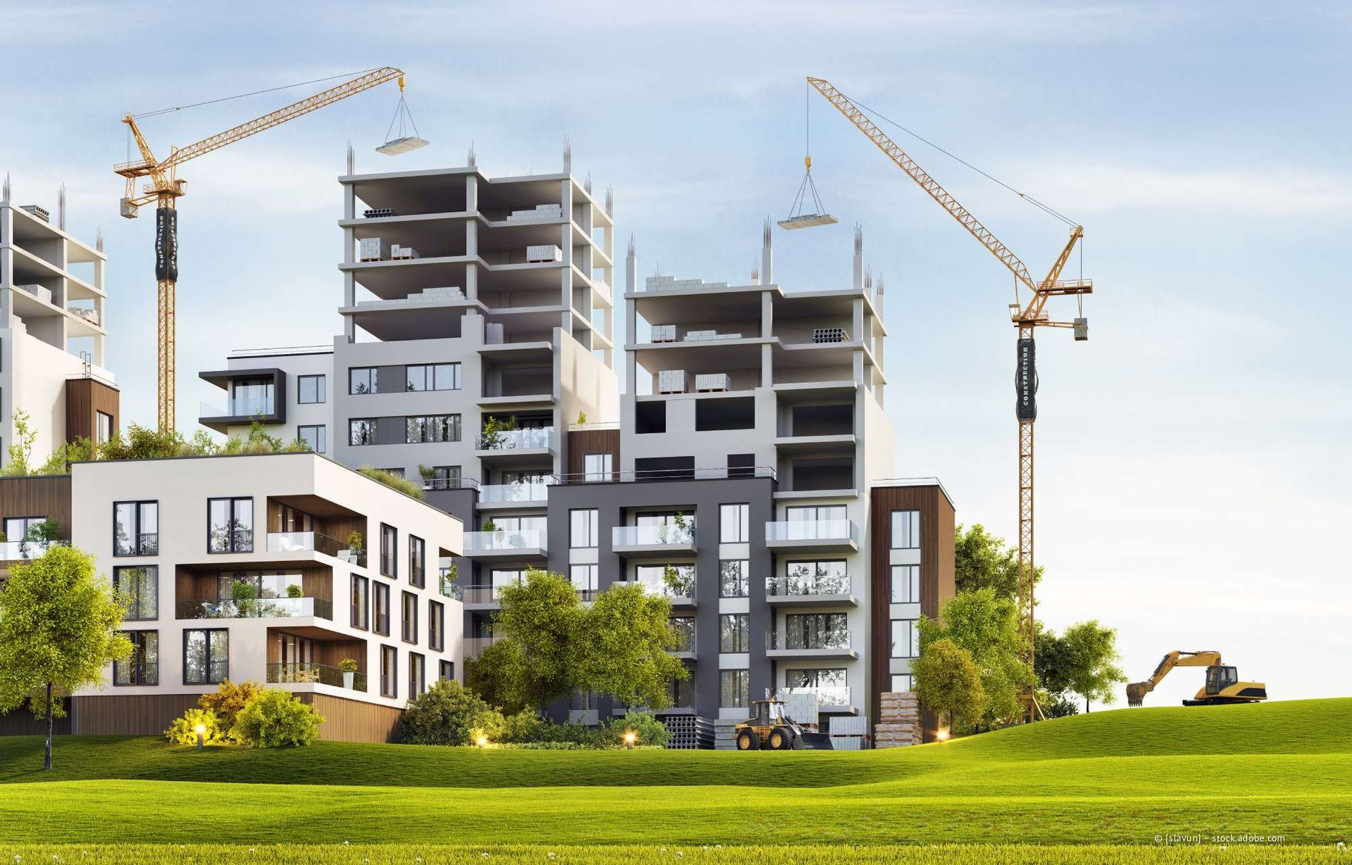 Im Bau befindliche Gebäude mit Kränen auf einer Wiese, Bäume und ein Bagger sind sichtbar.