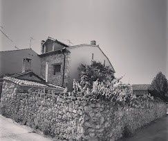 Casa de piedra con tejado de tejas y árboles junto a un camino estrecho en un entorno rural.