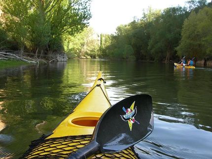 Kayak amarillo en el río, remo en foco, otros kayakistas en el fondo, árboles en las orillas.