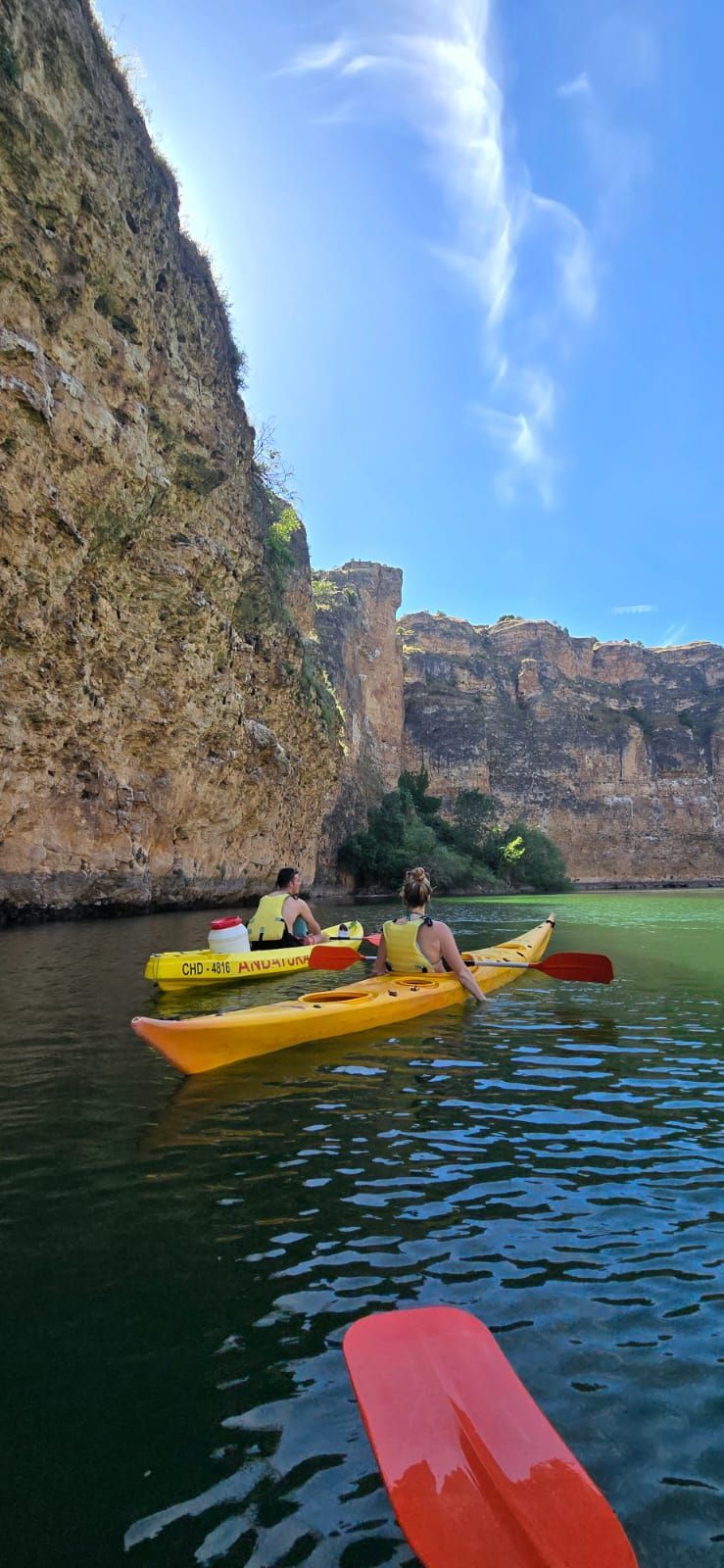 Dos personas haciendo kayak en un cañón con un acantilado rocoso, bajo un cielo azul.
