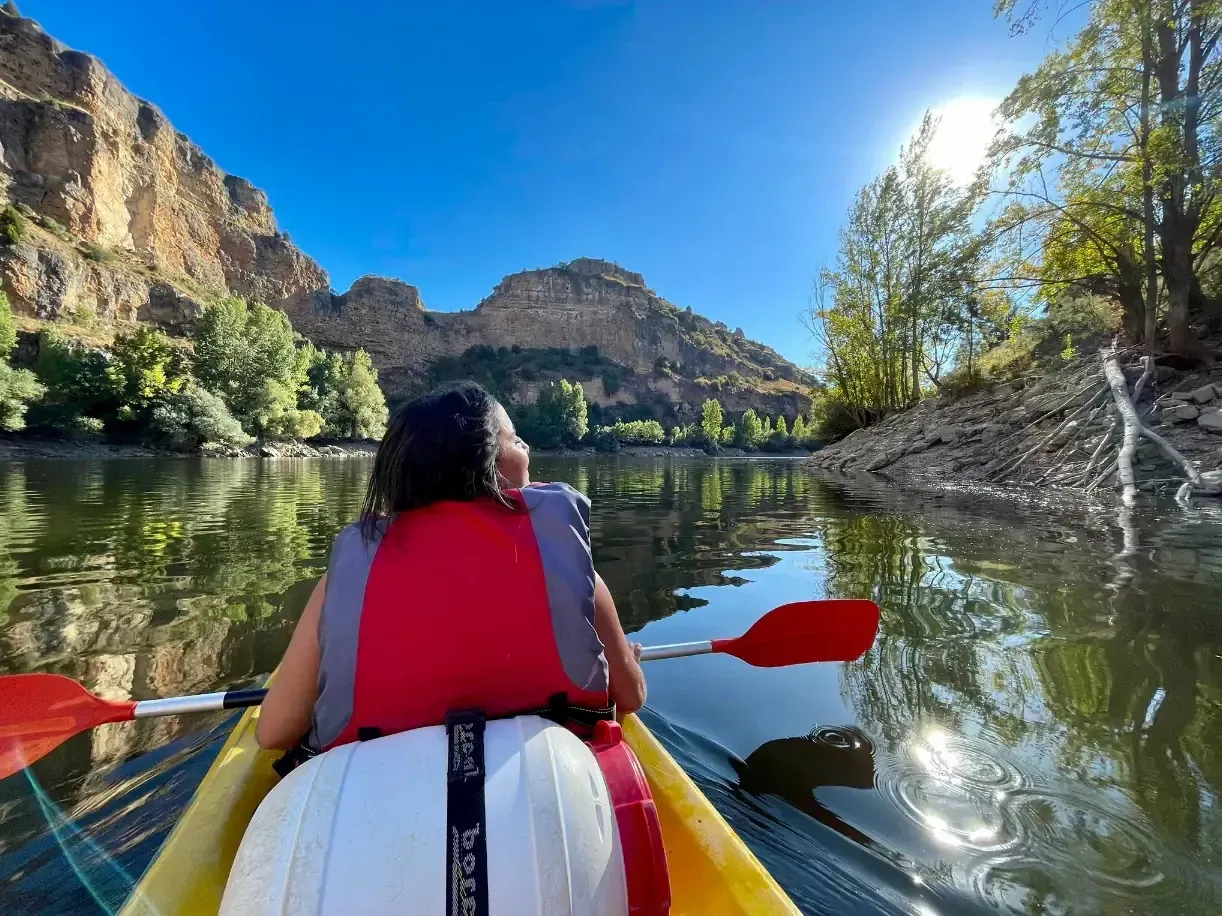Persona en kayak en un río, con acantilados y árboles al fondo bajo un cielo soleado.