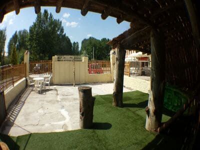 Patio exterior con césped artificial, soportes de madera y mesas. Día soleado con un edificio al fondo.