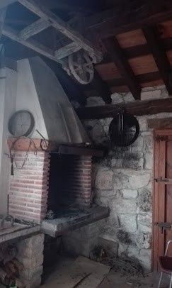 Antigua chimenea rústica de ladrillo con una gran campana, rueda colgante y vigas de madera en un interior con poca luz.