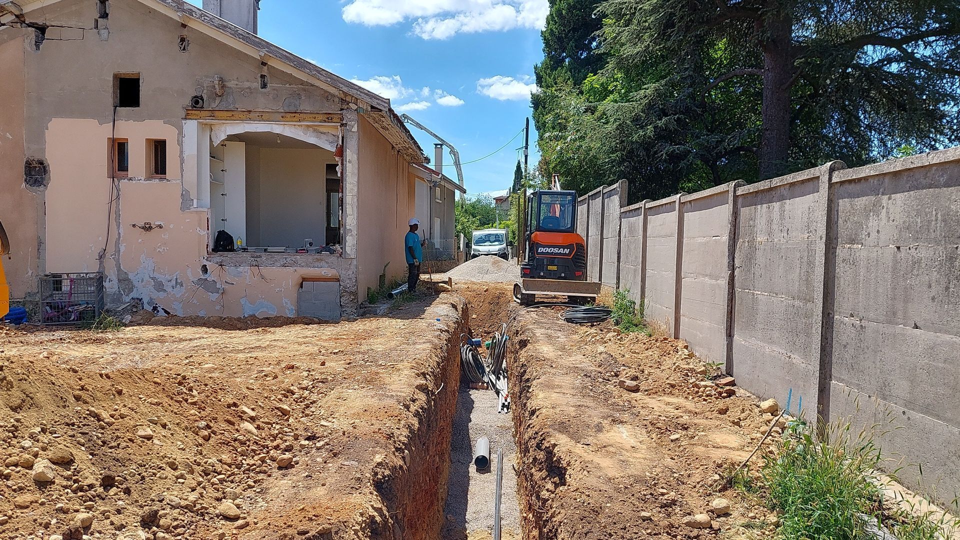 Chantier d'une tranchée creusée entre une maison et un mur en béton ; mini-excavatrice et ouvrier.