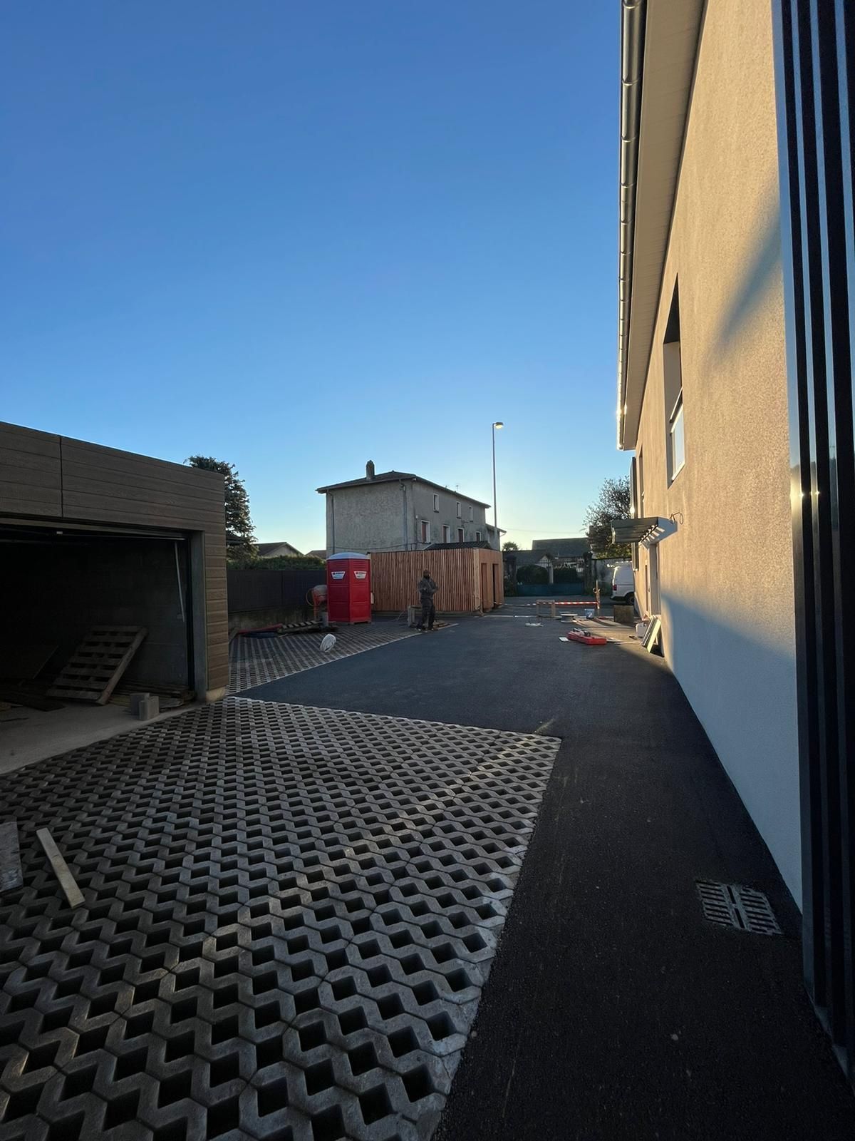 Allée asphaltée avec garage en construction, toilettes portatives et bâtiment par temps clair.