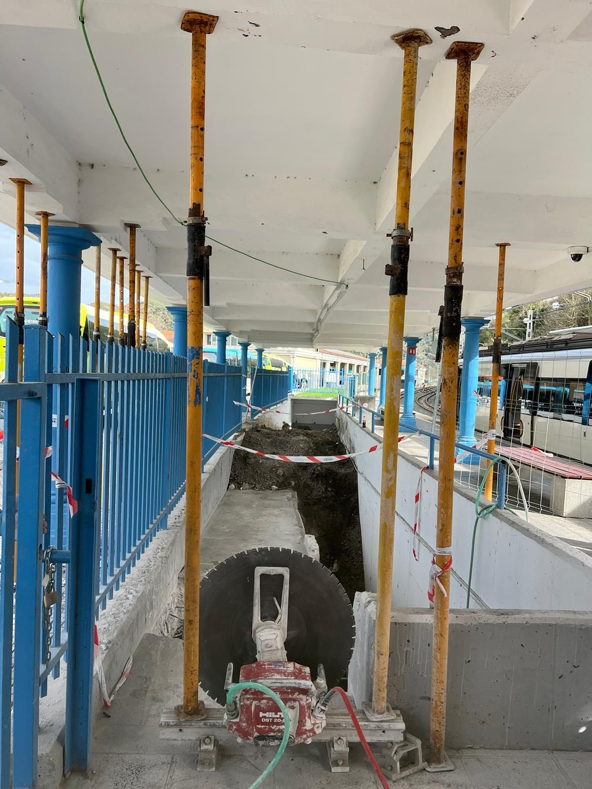 Obra en construcción con una perforadora roja en una zanja bajo andamios y postes de soporte azules.