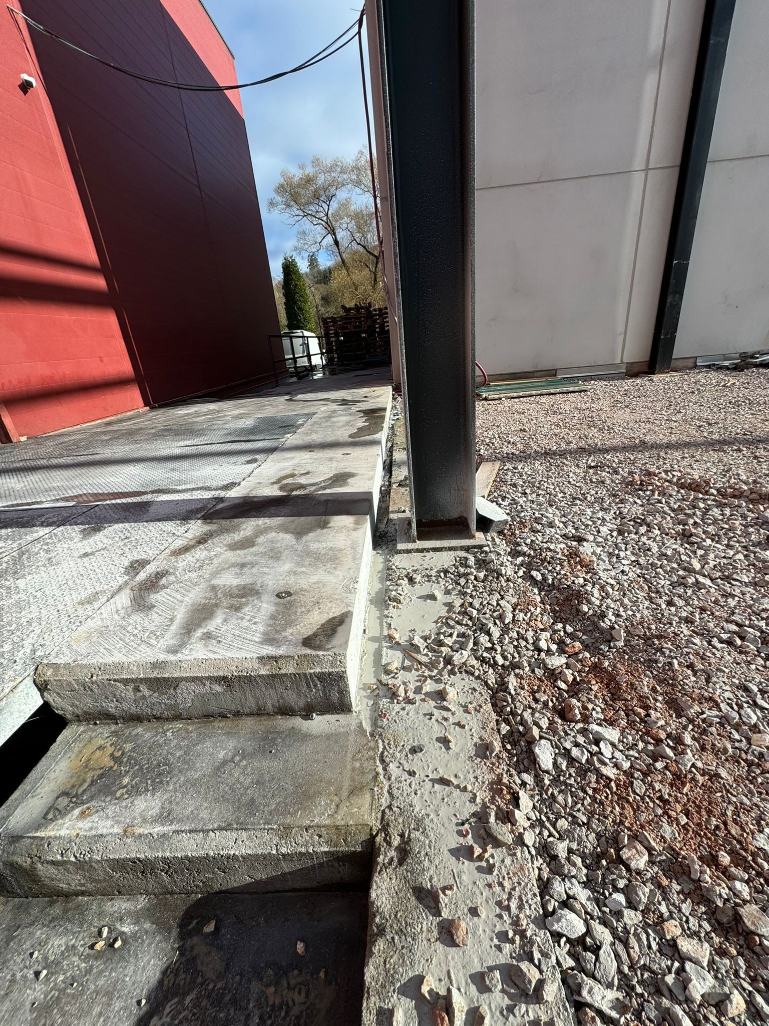 Escalones de piedra empinados junto a un poste negro, entre edificios rojos y blancos, con suelo de grava y árboles al fondo.