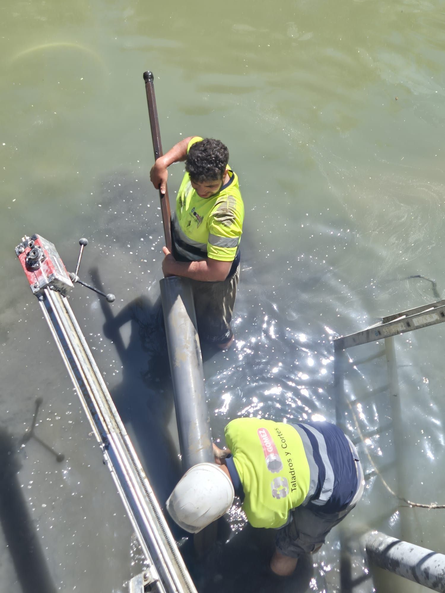 Dos trabajadores con chalecos de seguridad permanecen de pie en aguas turbias junto a una estructura metálica, manipulando un poste.