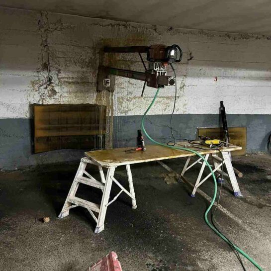 Taller con banco de trabajo, taladro de columna y herramientas en un garaje de hormigón con poca luz.