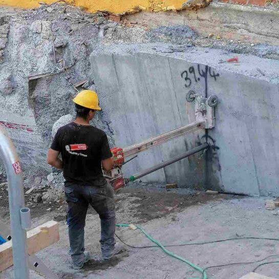 Un trabajador con casco amarillo utiliza una herramienta en un muro de hormigón en una obra de construcción.