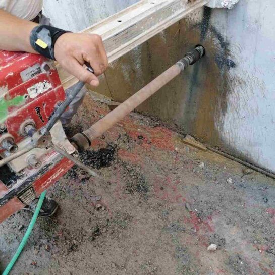 Un trabajador taladra una pared con una broca de núcleo rojo, generando polvo junto a un piso de concreto y una tubería.