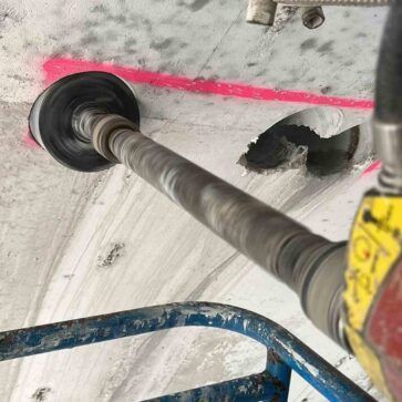Taladro cortando un agujero circular en un techo de hormigón, con línea de tiza rosa y andamios azules visibles.