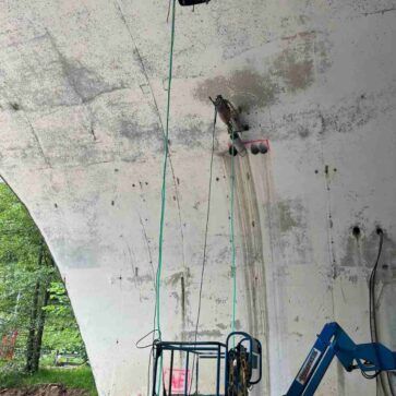 Ascensor azul bajo un arco de hormigón con cuerdas colgantes, en una zona boscosa.