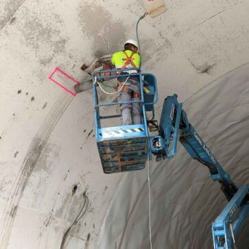 Un trabajador en una plataforma elevadora azul limpia el techo de un túnel de hormigón con una herramienta de mango largo.